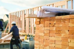 A closeup of stacks of 2x4 boards at a construction site, with a roll of blueprints sitting on top. Two construction workers and a building frame can be seen in the background. Horizontal shot.
