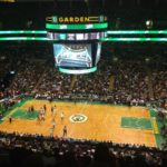 Players for the Celtics and Spurs basketball teams stand underneath the TD Garden's large video projectors, decorated with TD Bank logos, during a 2014 game with a full audience in the stands.