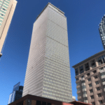 A photo of the Prudential Center skyscraper taken from the sidewalk, with the tower's shape elongated vertically against a cloudless blue sky.