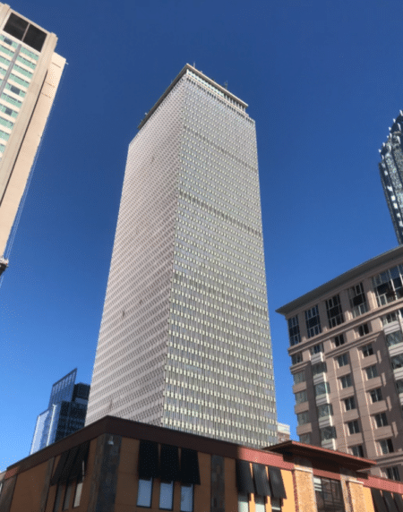 A photo of the Prudential Center skyscraper taken from the sidewalk, with the tower's shape elongated vertically against a cloudless blue sky.