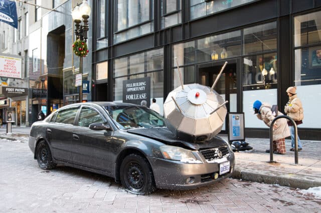 A Nissan Altima sits by the curb on Washington Street with a round, metal satellite on its hood. The satellite has crushed the car's hood in and popped a front tire. A pedestrian standing next to it has bent down to examine the sculpture more closely. 