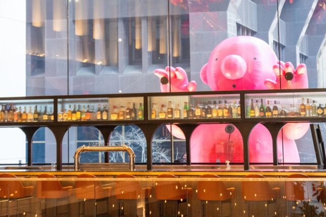 A roughly 12-foot-tall inflatable, pink,. bald cartoon man peeks into floor-to-ceiling windows of a bar in a downtown Boston skyscraper.