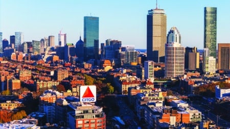 An aerial view of the Boston skyline featuring the iconic CITGO sign. The image captures the city's diverse architecture, including skyscrapers, historic buildings, and the Charles River. The CITGO sign stands out prominently against the backdrop of the cityscape.