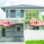 one hand holds out a stack of dollar bills from the left, a second hand holds out a model of a house from the right. A bungalow on a residential side street is behind them.