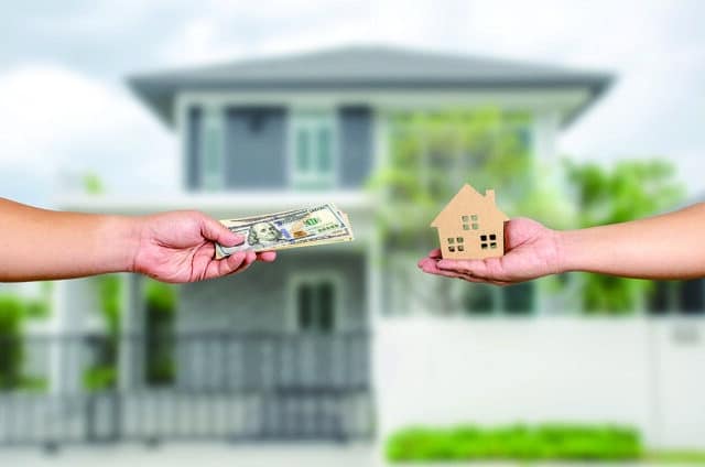 one hand holds out a stack of dollar bills from the left, a second hand holds out a model of a house from the right. A bungalow on a residential side street is behind them.