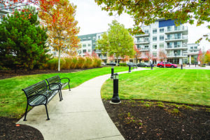 Couple of empty metal benches along a paved path lined with lamps in a park surrounded by modern apartment blocks on a cloudy autumn morning. Boston, MA, USA.